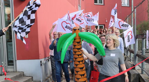 Eine Zielflagge links im Bild. Eine blonde Frau mit einer großen Gummi-Palme tritt über die Ziellinie (rotes Band) Hinter hier Teilnehmende im weißen AWO Flaggen.