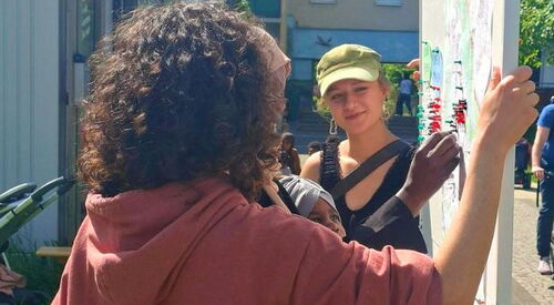 2 junge Menschen halten ein Pinntafel, währen eine Person einen Pin anpinnt. Draußen, Sonne scheint.