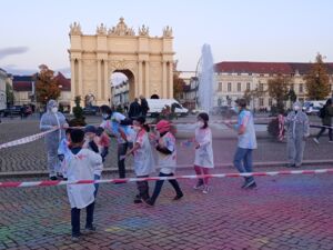 Der Luisenplatz mit Springbrunnen und brandenburger Tor im Hintergrund. Ein Absperrband wird von Menschen in Schutzanzügen / Masken gehalten sodass ein Rechteck umspannt ist, darin Mädchen in weißen Shirts bunt befleckt, auch der Steine / sind kunterbunt.